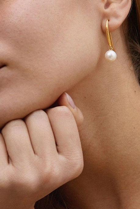 Close-up of a woman wearing a gold hoop earring with a pearl, against a neutral background.